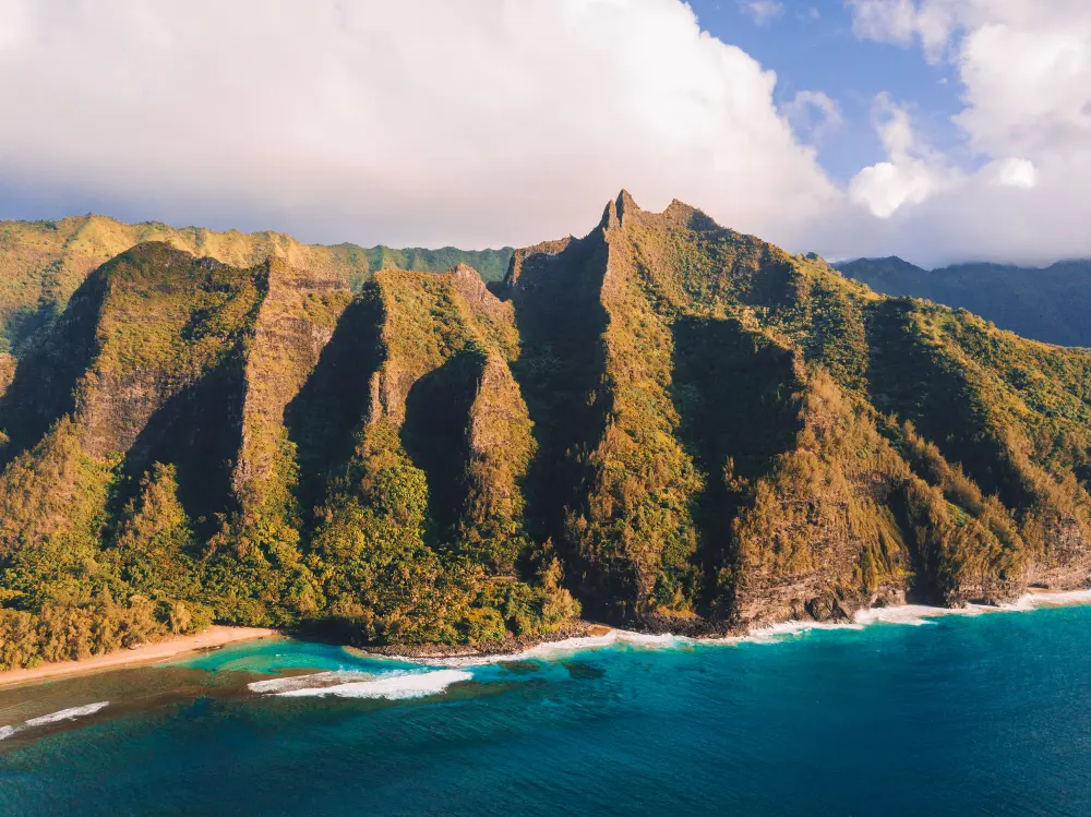 Aerial view of a Hawaiian beach with a commercial airplane flying overhead, illustrating how much are round trip plane tickets to Hawaii in 2026