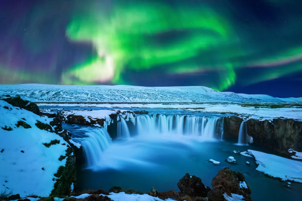 A stunning view of a glowing blue ice cave and the Northern Lights over a snowy mountain range, showing the winter beauty of how to plan a trip to Iceland.
